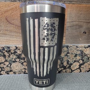 Pode incluir: Caneca isolada preta com aro e tampa prateados. A caneca apresenta um design de bandeira americana desgastada com os n&uacute;meros "45" e "47" no campo de estrelas da bandeira. A marca "YETI" &eacute; exibida na parte inferior.