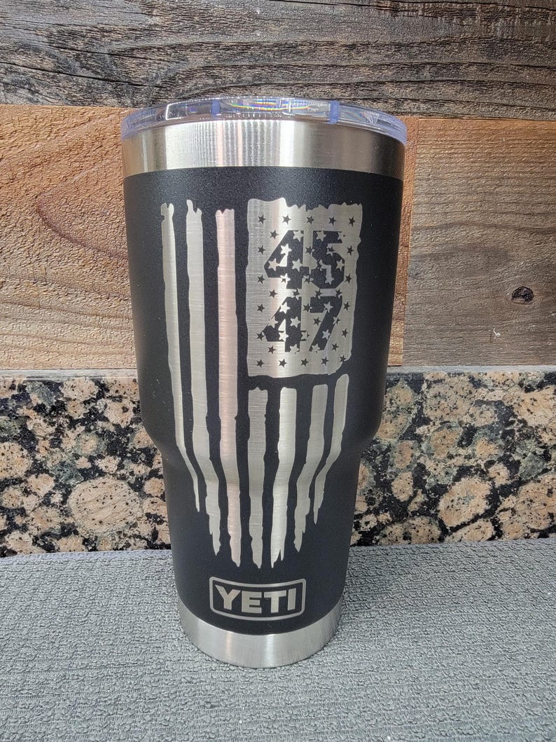 Pode incluir: Caneca isolada preta com aro e tampa prateados. A caneca apresenta um design de bandeira americana desgastada com os n&uacute;meros "45" e "47" no campo de estrelas da bandeira. A marca "YETI" &eacute; exibida na parte inferior.