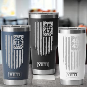 Pode incluir: Tr&ecirc;s canecas isoladas em azul marinho, preto e branco, cada uma com um design de bandeira americana desgastada e os n&uacute;meros "45 47" em um ret&acirc;ngulo cheio de estrelas. As canecas t&ecirc;m uma borda prateada e o logotipo "YETI".