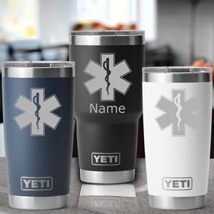 May include: Three stainless steel YETI tumblers with a medical symbol and the word "Name" on the black tumbler. The tumblers are in navy blue, black, and white.