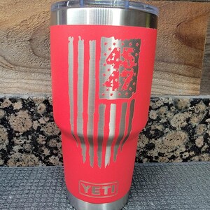 Pode incluir: Um copo isolado vermelho com tampa e base prateadas. O copo apresenta um design de bandeira americana desgastada com os n&uacute;meros "45" e "47" gravados na bandeira. A marca "YETI" &eacute; vis&iacute;vel na parte inferior.