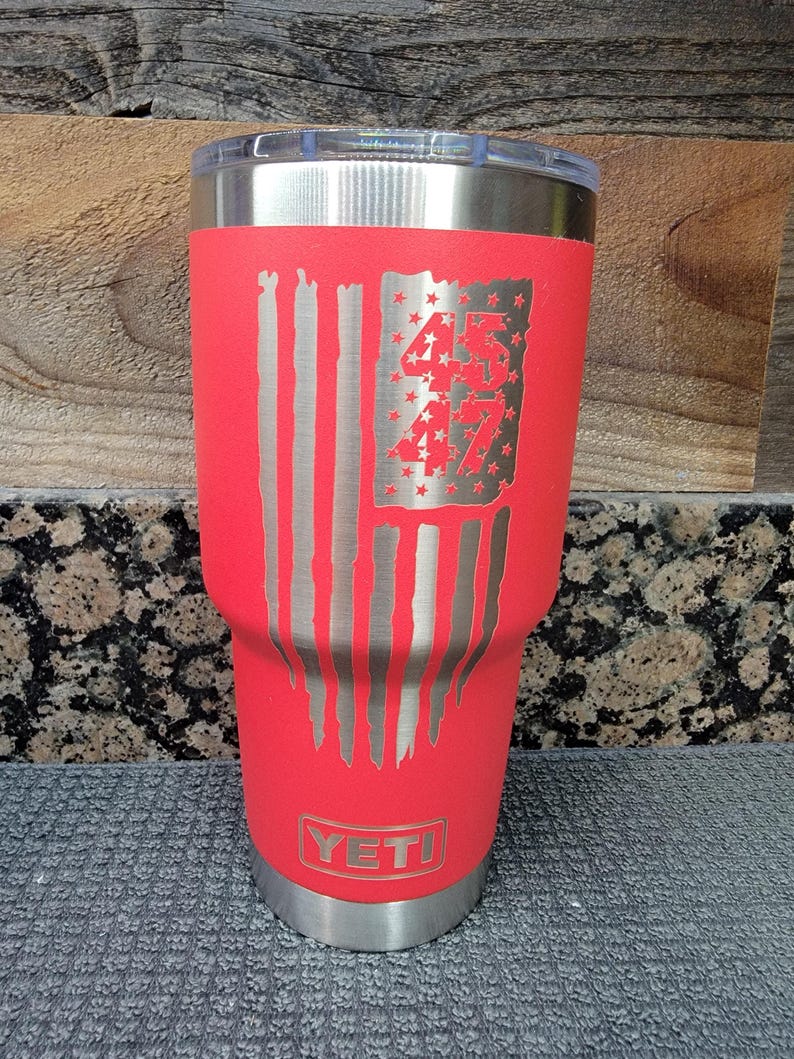 Pode incluir: Um copo isolado vermelho com tampa e base prateadas. O copo apresenta um design de bandeira americana desgastada com os n&uacute;meros "45" e "47" gravados na bandeira. A marca "YETI" &eacute; vis&iacute;vel na parte inferior.
