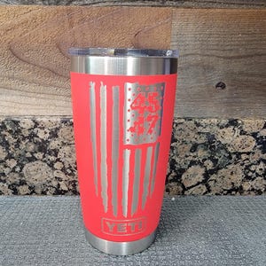 Pode incluir: Um copo isolado vermelho com tampa e base prateadas. O copo apresenta um design de bandeira americana gravado em prata com o n&uacute;mero "45" nas estrelas. A marca "YETI" &eacute; vis&iacute;vel na parte inferior.