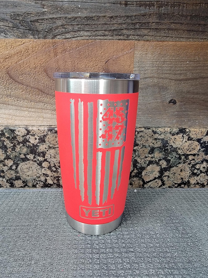 Pode incluir: Um copo isolado vermelho com tampa e base prateadas. O copo apresenta um design de bandeira americana gravado em prata com o n&uacute;mero "45" nas estrelas. A marca "YETI" &eacute; vis&iacute;vel na parte inferior.