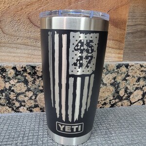 Pode incluir: Caneca isolada preta com tampa transparente. A caneca apresenta um design de bandeira americana desgastada com os n&uacute;meros "45" e "47" no campo de estrelas da bandeira. A marca "YETI" &eacute; exibida na parte inferior.