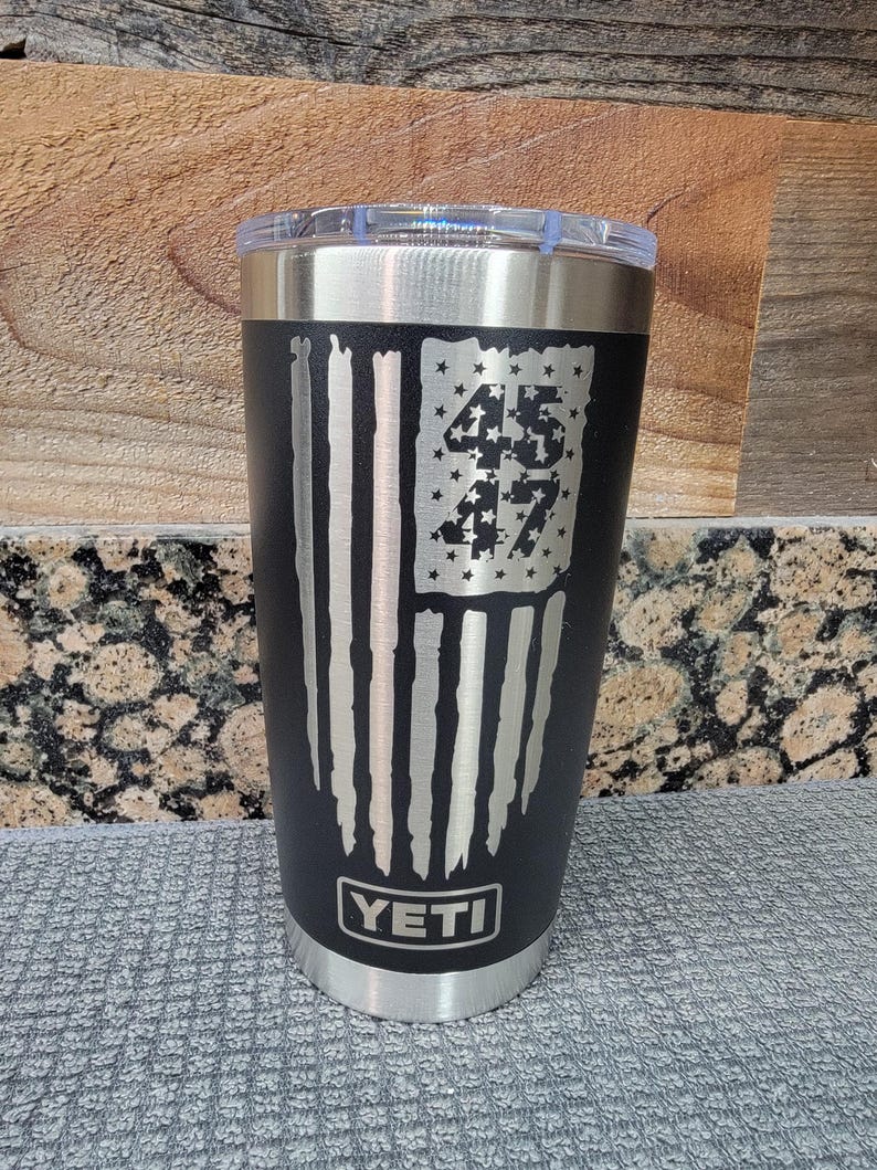 Pode incluir: Caneca isolada preta com tampa transparente. A caneca apresenta um design de bandeira americana desgastada com os n&uacute;meros "45" e "47" no campo de estrelas da bandeira. A marca "YETI" &eacute; exibida na parte inferior.