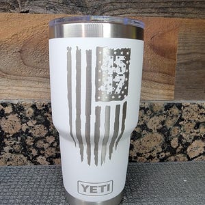Pode incluir: Um copo Yeti branco com tampa e base prateadas. O copo apresenta um design de bandeira americana desgastada com o n&uacute;mero "45" no campo de estrelas. O logotipo Yeti &eacute; exibido na parte inferior.
