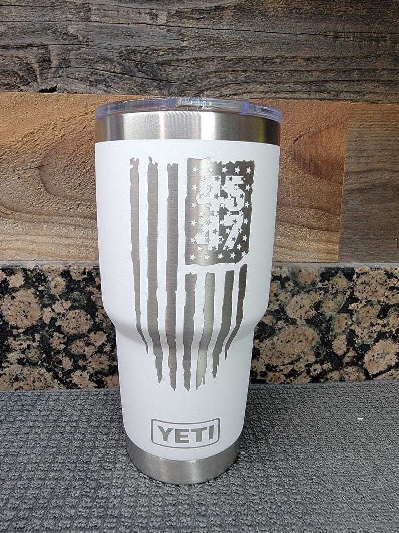 Pode incluir: Um copo Yeti branco com tampa e base prateadas. O copo apresenta um design de bandeira americana desgastada com o n&uacute;mero "45" no campo de estrelas. O logotipo Yeti &eacute; exibido na parte inferior.