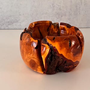 May include: A small, round wooden bowl crafted from a single piece of wood with a polished, glossy finish. The bowl features deep, natural cracks and dark, textured bark edges that contrast with the vibrant, warm orange-brown wood grain.