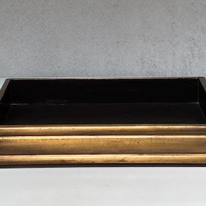 May include: A rectangular tray featuring a metallic gold finish on the exterior frame with a stepped, tiered edge design. The interior base and side walls of the tray are finished in a solid matte black color, creating a contrast with the gold border.