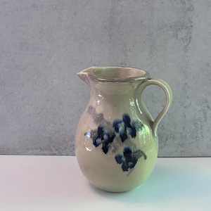 May include: A cream-colored ceramic pitcher with a rounded body, a curved handle, and a pouring spout. The exterior features a hand-painted floral design in shades of dark blue and grey, with a speckled texture across the glazed surface.