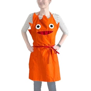 May include: Orange apron with a red mouth and two white eyes. The apron has a red tie.