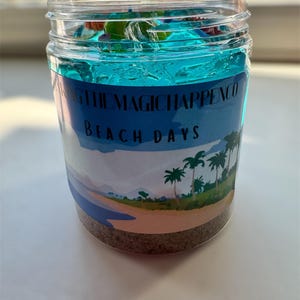 Puede incluir: Un tarro de vidrio transparente lleno de slime azul y arena. El tarro tiene una etiqueta que dice "Making the Magic Happen Co. Beach Days".