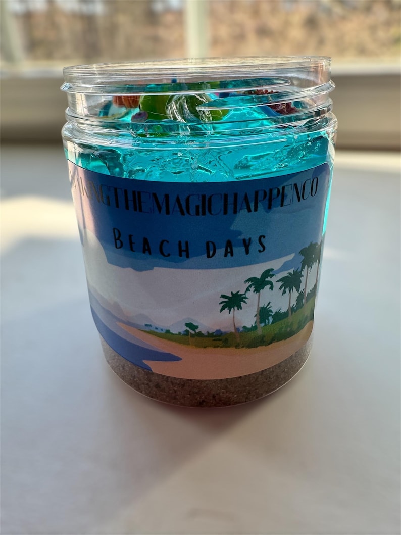Puede incluir: Un tarro de vidrio transparente lleno de slime azul y arena. El tarro tiene una etiqueta que dice "Making the Magic Happen Co. Beach Days".