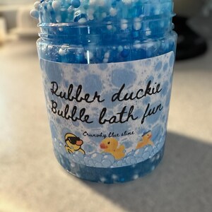 Op de afbeelding: Een blauwe slime met witte en iriserende kralen in een glazen pot. De pot heeft een etiket met de tekst "Rubber duckie bubble bath fun" en "Crunchy blue slime".