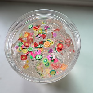 Pode incluir: Um slime transparente e claro em um recipiente redondo de plástico transparente. O slime é preenchido com amuletos coloridos em forma de fruta, incluindo fatias de melancia, laranja, morango e kiwi. O recipiente está em uma superfície branca.