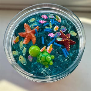 Puede incluir: Un tarro transparente lleno de slime azul, que contiene varias criaturas marinas de pl&aacute;stico de colores, incluyendo una tortuga, una estrella de mar y peces. El slime es texturizado y se asemeja al agua del mar.