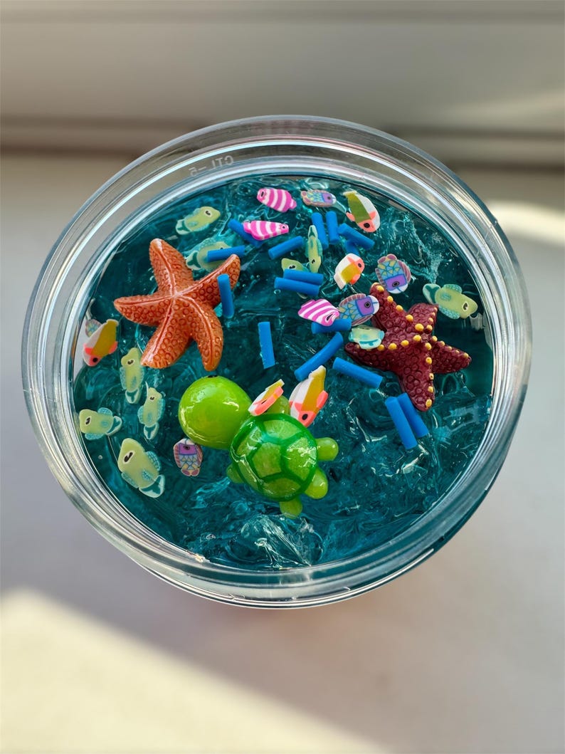 Puede incluir: Un tarro transparente lleno de slime azul, que contiene varias criaturas marinas de pl&aacute;stico de colores, incluyendo una tortuga, una estrella de mar y peces. El slime es texturizado y se asemeja al agua del mar.