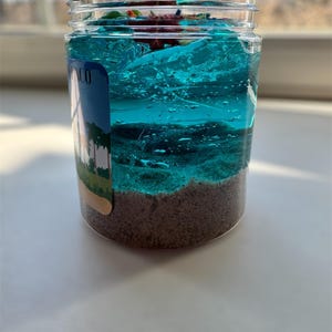 Puede incluir: Un tarro de vidrio transparente con slime azul y verde en capas. La slime est&aacute; cubierta de arena y una peque&ntilde;a pegatina de palmera. El tarro tiene una etiqueta que dice "MCO".