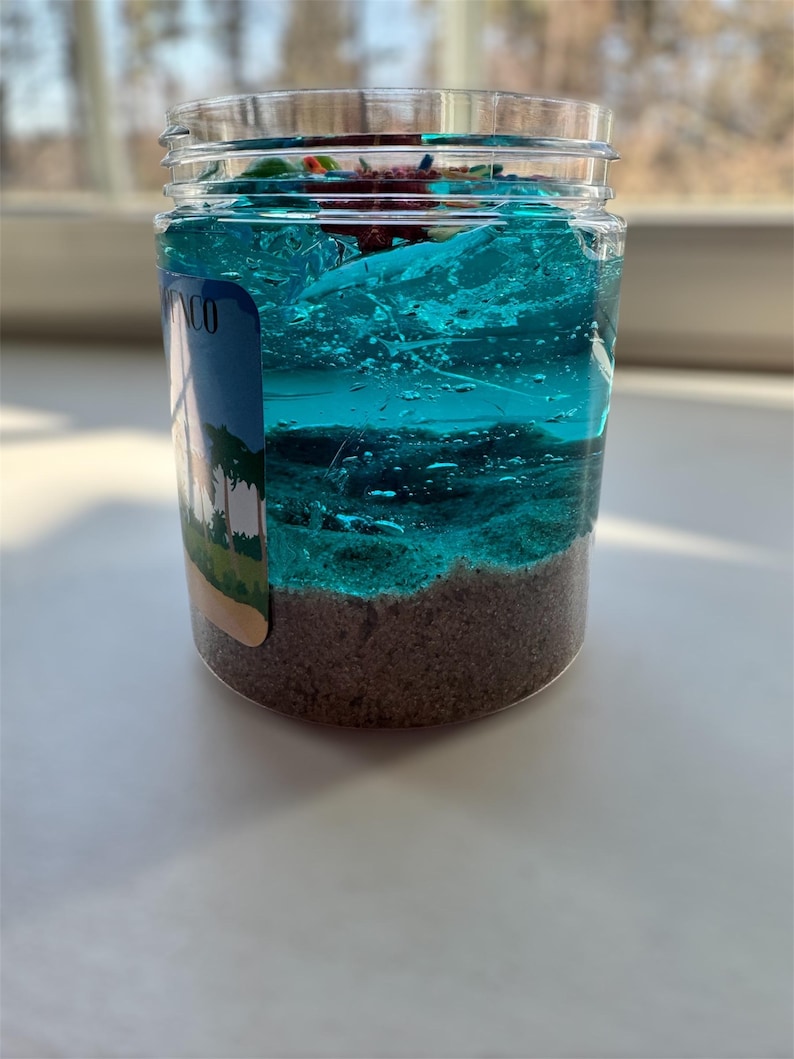 Puede incluir: Un tarro de vidrio transparente con slime azul y verde en capas. La slime est&aacute; cubierta de arena y una peque&ntilde;a pegatina de palmera. El tarro tiene una etiqueta que dice "MCO".