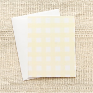 May include: A blank white card with a yellow and white gingham pattern on the front. The card is sitting on a beige textured surface.