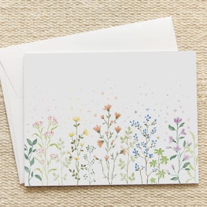 May include: A white stationery set featuring a floral watercolor design. The card displays a variety of colorful wildflowers in shades of pink, yellow, orange, blue, and purple. An envelope is included.