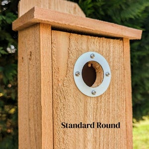 May include: A wooden birdhouse with a natural finish. It features a round entry hole with a white metal trim and the text "Standard Round" below the hole. The birdhouse has a pitched roof and is constructed from wooden planks.