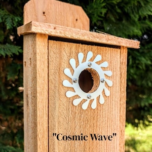 May include: A wooden birdhouse with a decorative white metal sunburst around the entry hole. The birdhouse is made of light brown wood and has a small roof. The text "Cosmic Wave" is printed on the front.