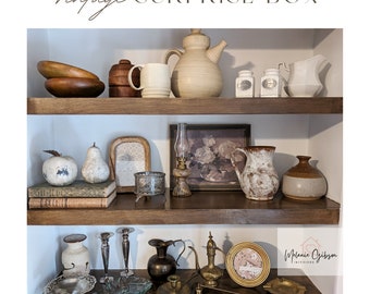 May include: A vintage surprise box filled with various items, including a teapot, mugs, bowls, a lamp, a pitcher, and a vase. The items are arranged on two wooden shelves. The text "Vintage Surprise Box" is written in cursive at the top of the image. The text "Melanie Gibson Interiors" is written in the bottom right corner of the image.