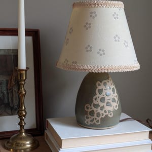 May include: A decorative table lamp with a cream lampshade featuring a floral pattern and a light pink trim. The base is a muted green with a white floral design. A brass candlestick and a framed picture are also visible.