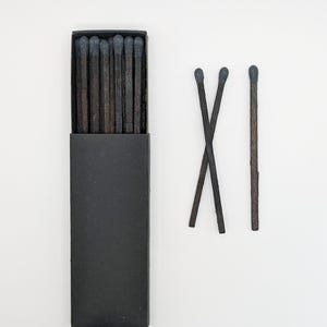 May include: A rectangular black matchbox containing several wooden matches with dark brown stems and charcoal gray heads. Two additional matches with dark brown stems and charcoal gray heads are positioned to the right of the box, with one match placed vertically and two others crossed in an X shape.
