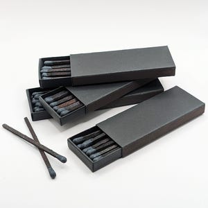 May include: Three rectangular black matchboxes with sliding trays containing wooden matches with dark charcoal-colored heads. Two loose matches with dark charcoal heads and dark brown wooden sticks are positioned in front of the boxes.