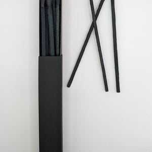 May include: A black matchbox containing dark grey matches, with several matches scattered nearby. The match heads are a darker shade of grey. The matches are set against a white background.