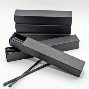 Puede incluir: Varias cajas de cerillas negras apiladas, una abierta que revela cerillas gris oscuro. Dos cerillas fuera de la caja, mostrando las cabezas grises. Las cajas son negro mate, sobre un fondo blanco.