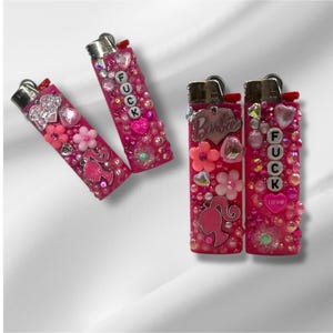 Puede incluir: Encendedores rosas decorados con cuentas, flores y abalorios. Un encendedor tiene la palabra "FUCK" escrita, mientras que otro presenta la palabra "Barbie" y una silueta. Los encendedores est&aacute;n adornados con cuentas rosas y transparentes.