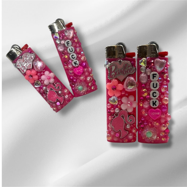 Puede incluir: Encendedores rosas decorados con cuentas, flores y abalorios. Un encendedor tiene la palabra "FUCK" escrita, mientras que otro presenta la palabra "Barbie" y una silueta. Los encendedores est&aacute;n adornados con cuentas rosas y transparentes.