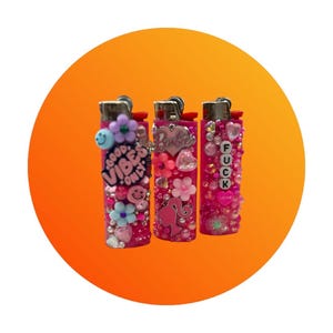 Puede incluir: Tres encendedores rosas decorados con pedrer&iacute;a, flores y abalorios. Un encendedor tiene el texto "GOOD VIBES ONLY", otro dice "Barbie" y el tercero dice "FUCK LOVE". Los encendedores est&aacute;n sobre un fondo naranja.