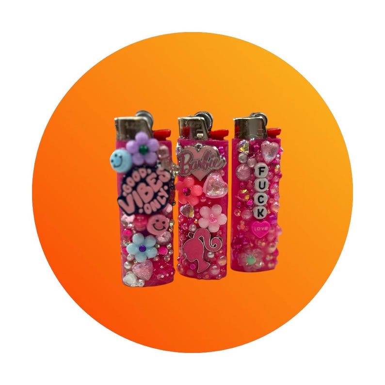 Puede incluir: Tres encendedores rosas decorados con pedrer&iacute;a, flores y abalorios. Un encendedor tiene el texto "GOOD VIBES ONLY", otro dice "Barbie" y el tercero dice "FUCK LOVE". Los encendedores est&aacute;n sobre un fondo naranja.