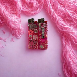 Puede incluir: Dos encendedores rosas decorados con adornos. Uno tiene la palabra "Barbie" y detalles florales. El otro tiene la palabra "FUCK" escrita con cuentas y un coraz&oacute;n que dice "LOVE".