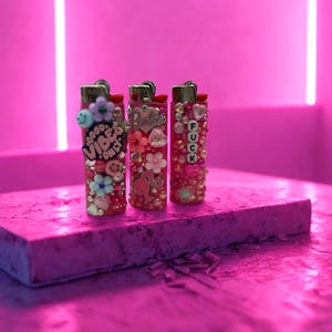 Puede incluir: Tres encendedores decorativos con adornos rosas y rojos, incluyendo flores, cuentas y dijes. Un encendedor tiene el texto "GOOD VIBES ONLY", otro tiene un dije de "Barbie", y el tercero tiene las letras "FUCK".