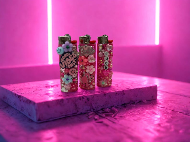 Puede incluir: Tres encendedores decorativos con adornos rosas y rojos, incluyendo flores, cuentas y dijes. Un encendedor tiene el texto "GOOD VIBES ONLY", otro tiene un dije de "Barbie", y el tercero tiene las letras "FUCK".