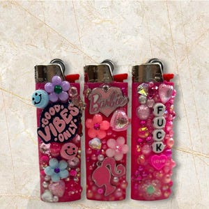 Puede incluir: Tres encendedores rosas decorados con varios adornos. Uno tiene el texto "GOOD VIBES ONLY" y una carita sonriente. Otro tiene el logo de "Barbie" y una silueta. El tercero tiene la palabra "FUCK" escrita con cuentas y un coraz&oacute;n.