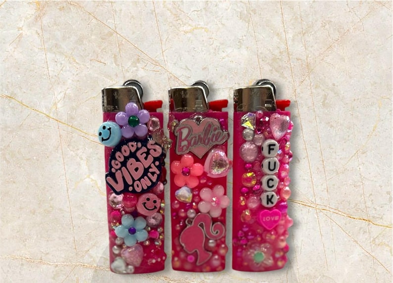 Puede incluir: Tres encendedores rosas decorados con varios adornos. Uno tiene el texto "GOOD VIBES ONLY" y una carita sonriente. Otro tiene el logo de "Barbie" y una silueta. El tercero tiene la palabra "FUCK" escrita con cuentas y un coraz&oacute;n.