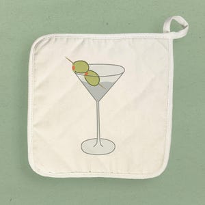 May include: A white quilted pot holder featuring a martini glass illustration. The martini glass is filled with a light gray liquid and garnished with two green olives on a toothpick. The pot holder has a loop for hanging.