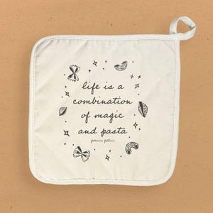 May include: White cotton pot holder with a black quote that reads "life is a combination of magic and pasta." The quote is attributed to Federico Fellini. The pot holder is decorated with black pasta shapes and stars.