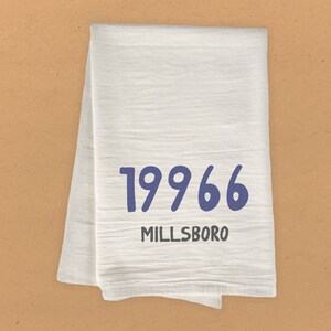 May include: White kitchen towel with the text "1996" in blue and "MILLSBORO" in black.