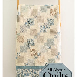 Könnte beinhalten: Ein Quilt mit einem Patchwork aus quadratischen Stoffblöcken in Creme-, Blau- und Brauntönen mit Blumen- und geometrischen Mustern. Der Quilt hat einen einfarbigen blauen Rand am unteren Ende und hängt auf einer Holzleiter. Text lautet Cheese & Crackers.