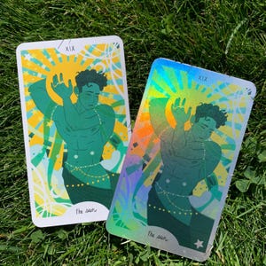 May include: Two tarot cards depicting a teal figure with raised arms, set against a yellow and blue sunburst. The cards are labelled "XIX" and "the sun". One card has a holographic finish.