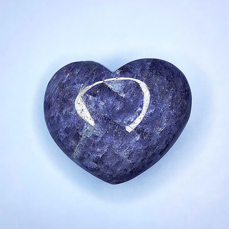 Puede incluir: Una piedra en forma de coraz&oacute;n en tonos azules y morados, con un dise&ntilde;o blanco curvado en su superficie. La piedra tiene un aspecto texturizado y est&aacute; sobre un fondo azul claro. Esta piedra decorativa podr&iacute;a usarse como pisapapeles o art&iacute;culo decorativo.