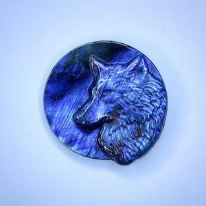 May include: A carved labradorite stone featuring a wolf's head profile. The sculpture is a deep blue color with iridescent flashes of purple and green. The wolf is set against a circular background.
